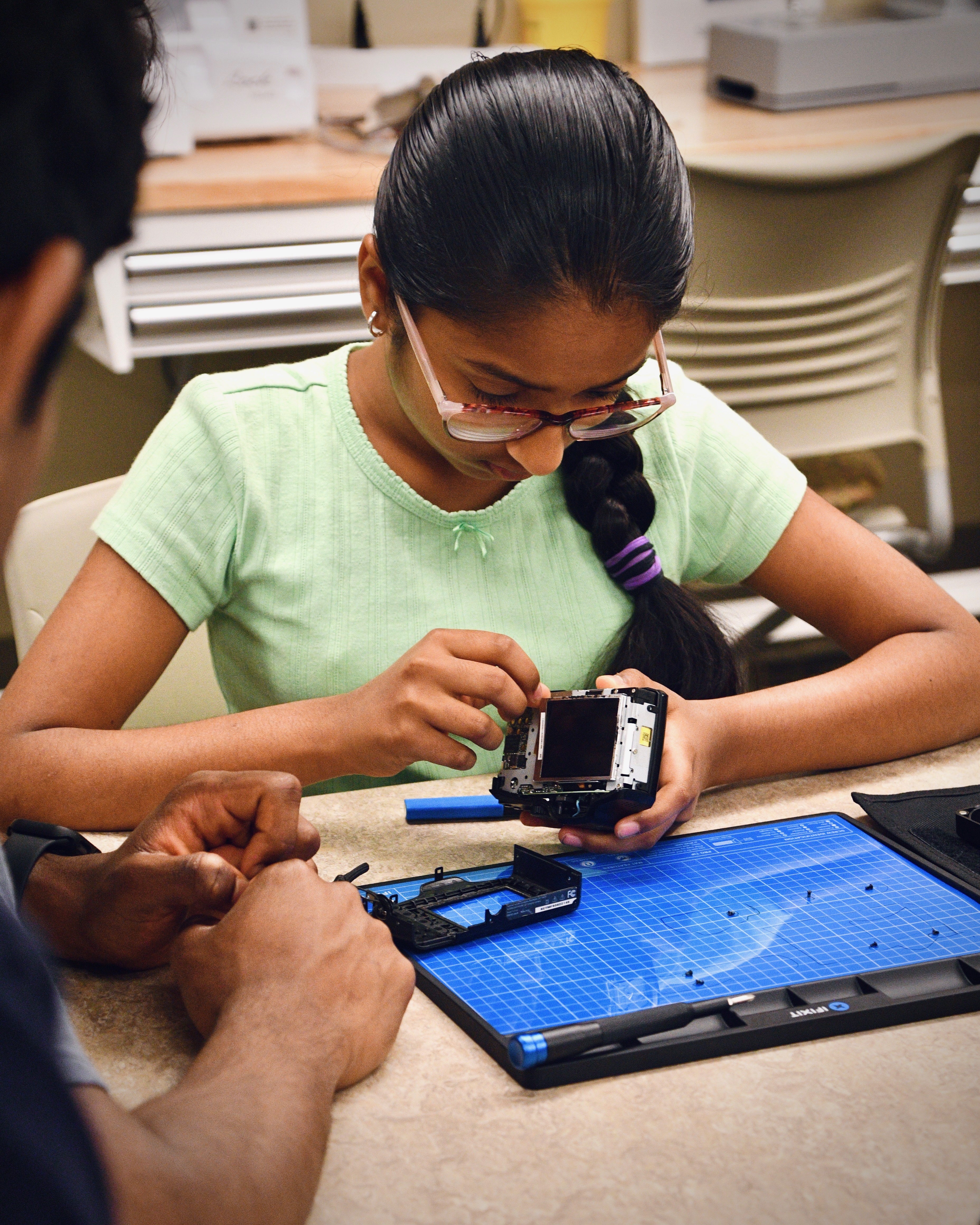 Student examining a disassembled camera over a FixMat at a repair workshop