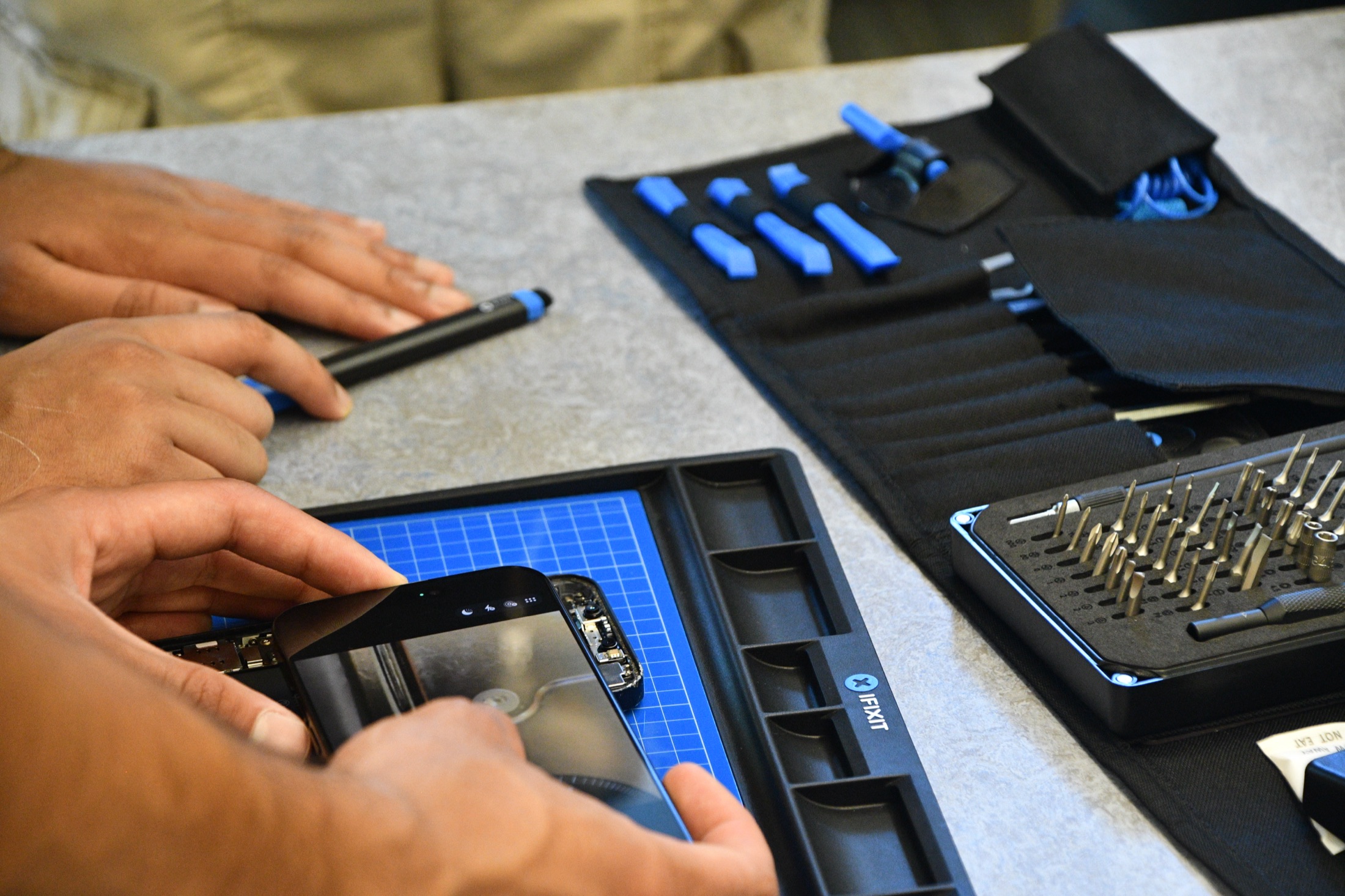 Overhead view of an iFixit Pro Tech Toolkit, bit driver set, and a phone being repaired on a blue FixMat